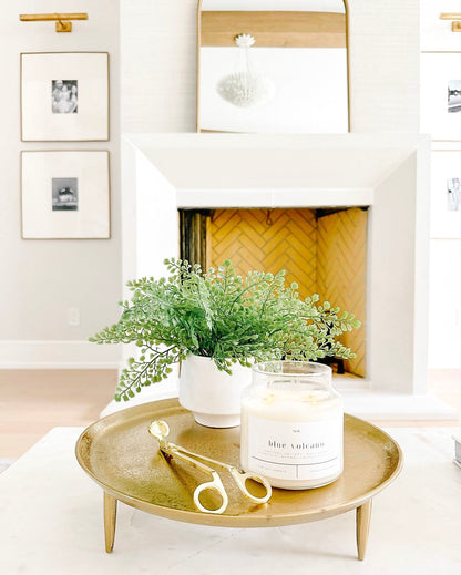 Blue Volcano 100% soy wax candle displayed on a gold tray alongside a green plant and wick trimmer, styled in a bright, modern living room. Hand-poured, eco-friendly candle with tropical fragrance notes of goji berry, pineapple, mango, and smoked vanilla, perfect for clean, non-toxic home decor.