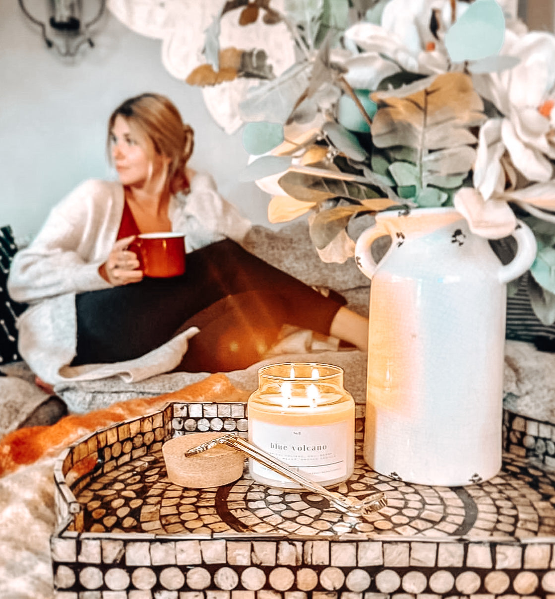 Blue Volcano 100% soy wax candle lit on a decorative tray with a cork lid and wick trimmer, styled in a cozy setting with a woman relaxing in the background. Hand-poured, eco-friendly candle with tropical fragrance notes of goji berry, pineapple, mango, and smoked vanilla, perfect for creating a warm, non-toxic ambiance.