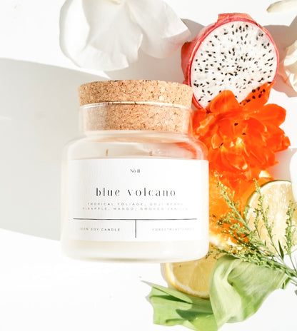 Blue Volcano 100% soy wax candle with a cork lid, styled with tropical fruits and vibrant flowers. Hand-poured, eco-friendly candle featuring notes of tropical foliage, goji berry, pineapple, mango, and smoked vanilla, perfect for a fresh, non-toxic home fragrance.